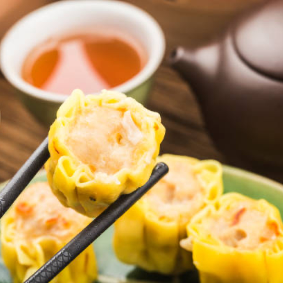 Chopsticks holding a yellow dim sum dumpling, with a teapot and teacup in the background.