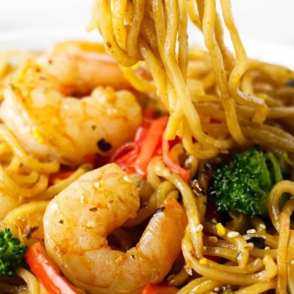 Close-up of shrimp and noodles with broccoli, red peppers, and sesame seeds being lifted.