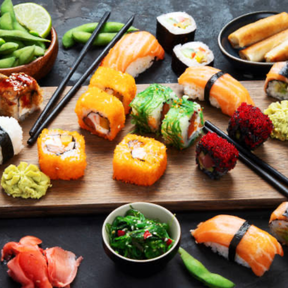 Assorted sushi rolls, nigiri, edamame, and spring rolls on a wooden board and dark surface.
