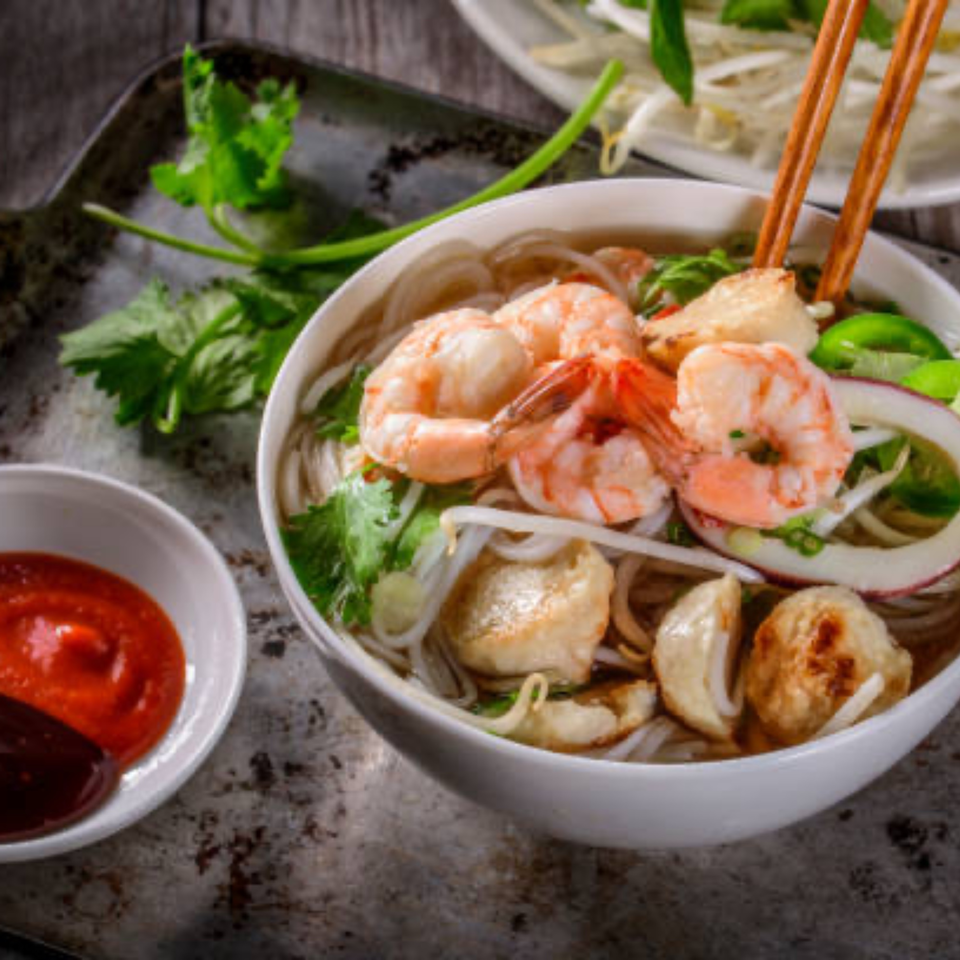 A bowl of Vietnamese pho with shrimp, noodles, and vegetables, with chopsticks and sauces.