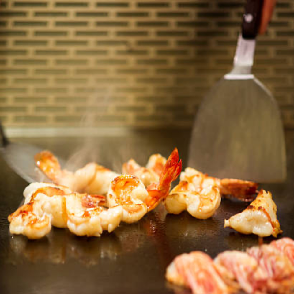 Shrimp and other ingredients cooking on a sizzling hibachi grill with spatulas.