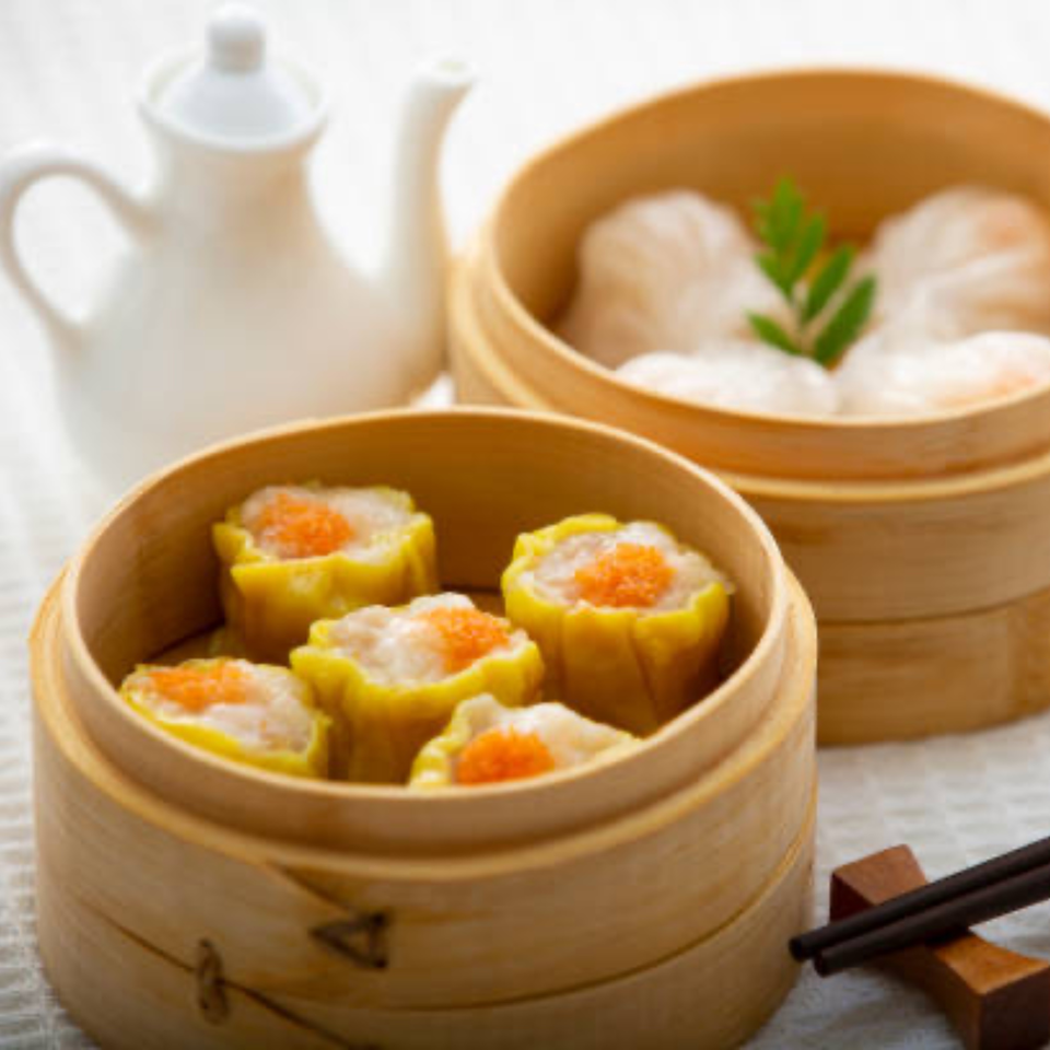 Steaming dim sum, including shumai and dumplings, in bamboo baskets with a teapot and chopsticks.