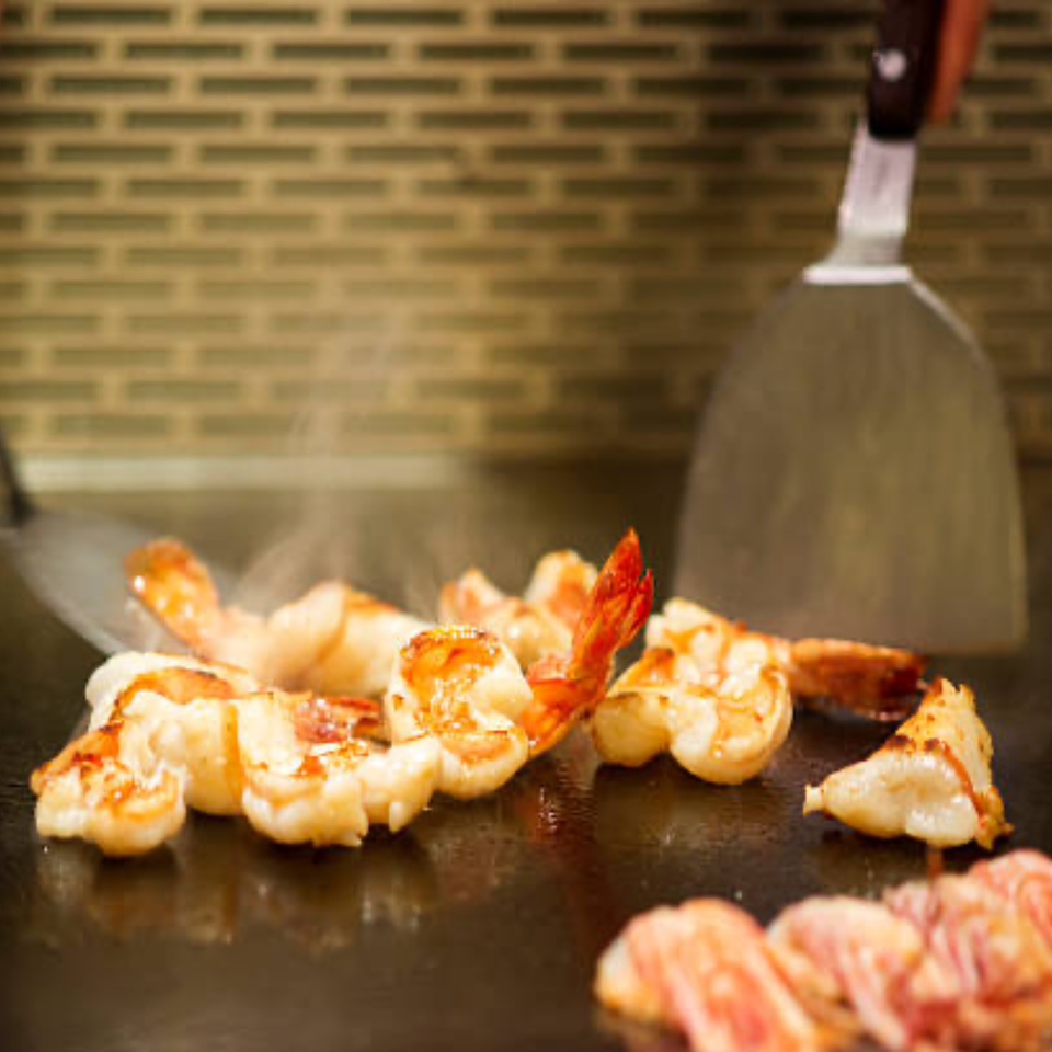 Shrimp and other seafood cooking on a sizzling hibachi grill with spatulas.