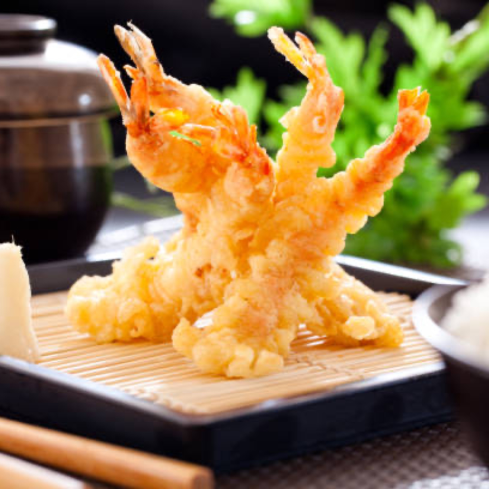 Golden-fried tempura shrimp on a bamboo mat, with a dark bowl and green foliage in the background.