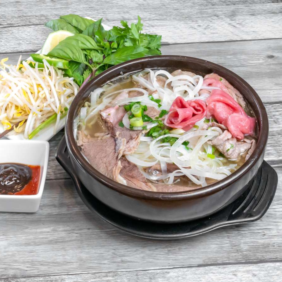 A bowl of Vietnamese pho with beef, noodles, and fresh herbs, bean sprouts, and sauces on the side.